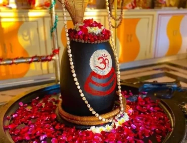 A black Shiva lingam adorned with a golden multi-hooded serpent, painted Om, rudraksha garland, fresh rose petals, and offerings in a temple setting.