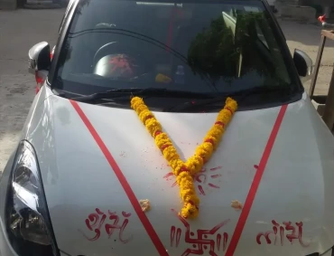 White car decorated with marigold garlands, red ribbons, and Hindu symbols for new-vehicle puja.