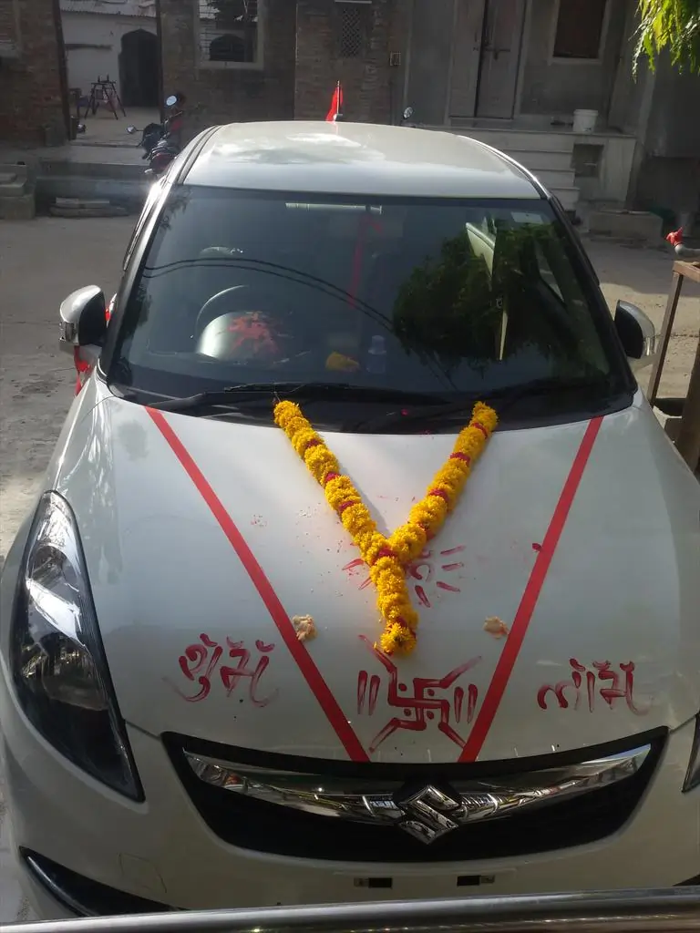 White car decorated with marigold garlands, red ribbons, and Hindu symbols for new-vehicle puja.