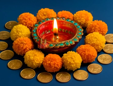 Lit red diya surrounded by turquoise-studded rim, marigold pom-poms, and arranged coins symbolizing Dhanteras prosperity.
