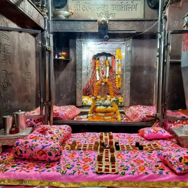 Sacred Omkareshwar Jyotirlinga sanctum with Shiva lingam, marigold garlands, silver vessels, embroidered pink cushions.
