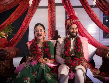 Smiling Hindu bride and groom in traditional attire sit under red drapes, wearing rose garlands
