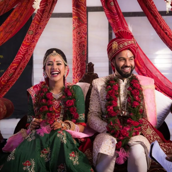 Smiling Hindu bride and groom in traditional attire sit under red drapes, wearing rose garlands