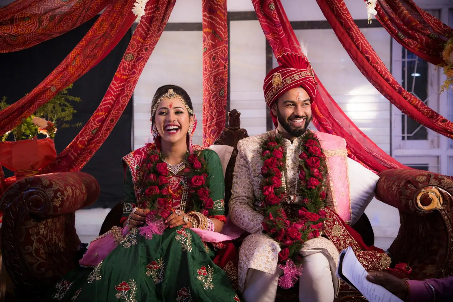 Smiling Hindu bride and groom in traditional attire sit under red drapes, wearing rose garlands